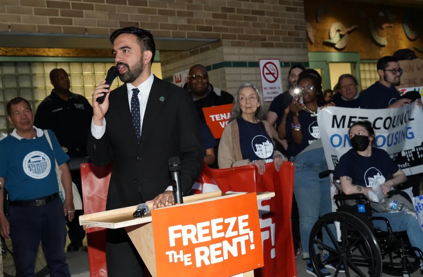 Zohran Mamdani speaking at a microphone behind a podium sign reading Freeze the Rent, with tenant organizers behind him